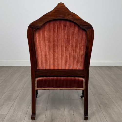 fig 3. A Victorian rosewood armchair featuring a dark timber frame, carved scroll detailing, rust red velvet upholstery, and turned front legs on castors.