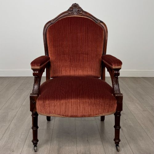 fig 7. A Victorian rosewood armchair featuring a dark timber frame, carved scroll detailing, rust red velvet upholstery, and turned front legs on castors.