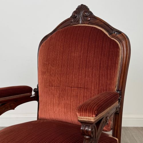 fig 8. A Victorian rosewood armchair featuring a dark timber frame, carved scroll detailing, rust red velvet upholstery, and turned front legs on castors.