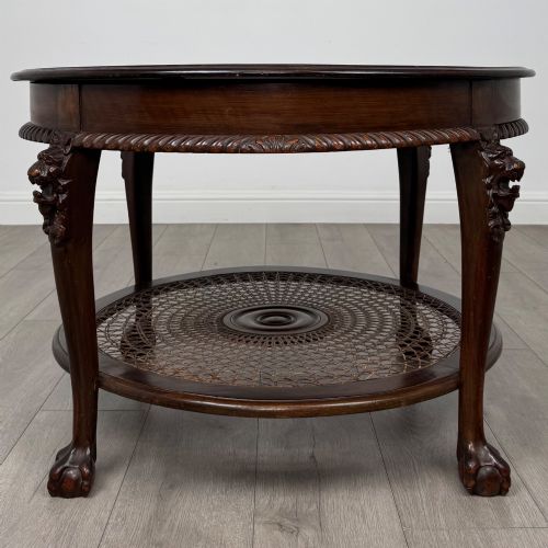 fig 2. A circular dark mahogany coffee table featuring carved lion heads atop cabriole legs ending in claw and ball feet, with a woven cane lower shelf.