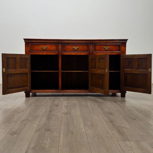 fig 1. A twentieth-century warm oak sideboard featuring three upper drawers with brass handles above three cupboard doors decorated with geometric moulded panels and exterior brass H-hinges.