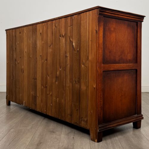 fig 6. A twentieth-century warm oak sideboard featuring three upper drawers with brass handles above three cupboard doors decorated with geometric moulded panels and exterior brass H-hinges.