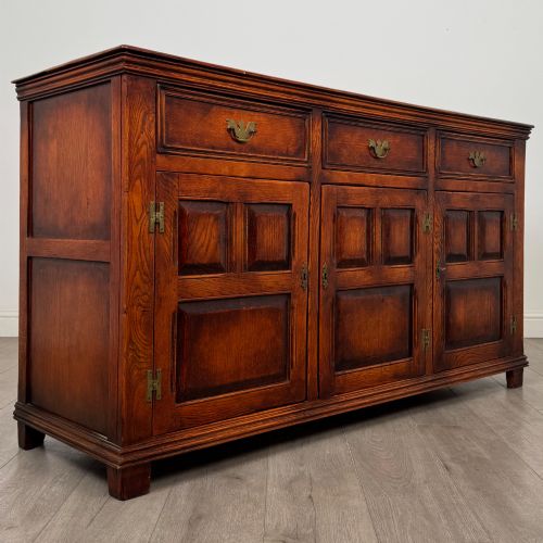 fig 8. A twentieth-century warm oak sideboard featuring three upper drawers with brass handles above three cupboard doors decorated with geometric moulded panels and exterior brass H-hinges.