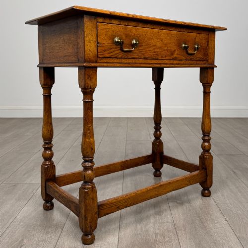 fig 7. A golden oak side table featuring a single drawer with two brass handles, turned legs, and an lower stretcher base.