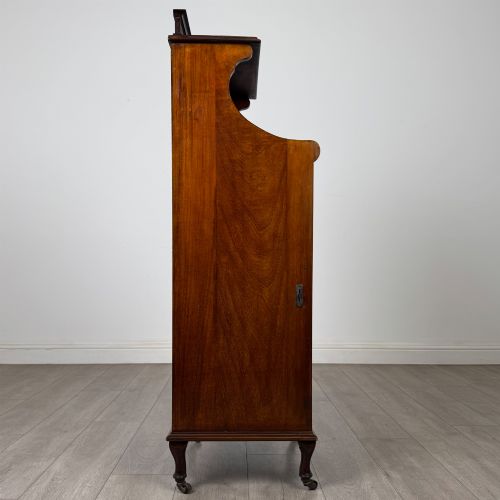 fig 7. An early 20th-century mahogany music cabinet with an upper mirrored shelf, a middle glazed drawer compartment, a lower moulded panel door, and brass handles on castors.