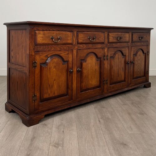 fig 8. A large medium brown oak sideboard featuring four upper drawers with brass handles above four cupboard doors with arched wood panels.