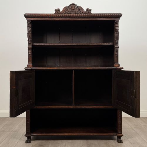 fig 1. A 19th-century dark oak carved cupboard featuring an open top shelf, carved lion cresting, relief-carved panel doors with figures, and an open lower base.