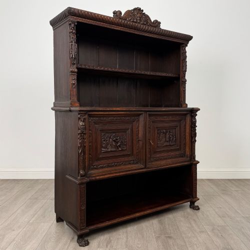 fig 8. A 19th-century dark oak carved cupboard featuring an open top shelf, carved lion cresting, relief-carved panel doors with figures, and an open lower base.