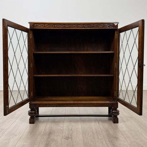 fig 1. An Edwardian dark oak display cabinet with double diamond-leaded glass doors, a carved scroll frieze along the top, interior wooden shelves, and bulbous turned legs connected by a stretcher.
