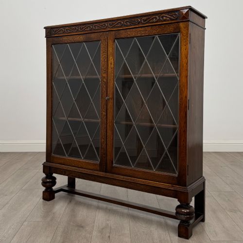 fig 2. An Edwardian dark oak display cabinet with double diamond-leaded glass doors, a carved scroll frieze along the top, interior wooden shelves, and bulbous turned legs connected by a stretcher.