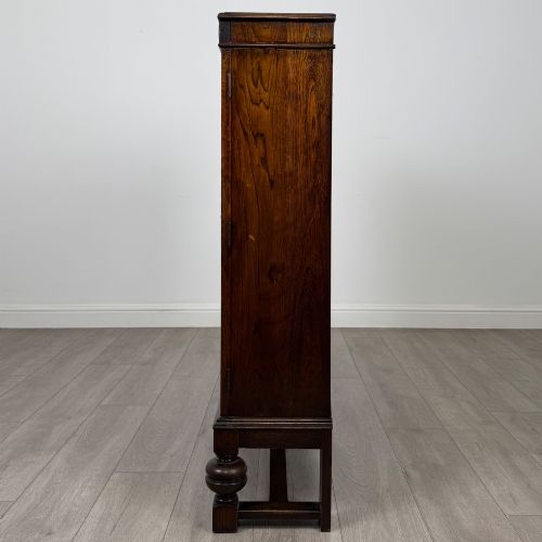 fig 3. An Edwardian dark oak display cabinet with double diamond-leaded glass doors, a carved scroll frieze along the top, interior wooden shelves, and bulbous turned legs connected by a stretcher.