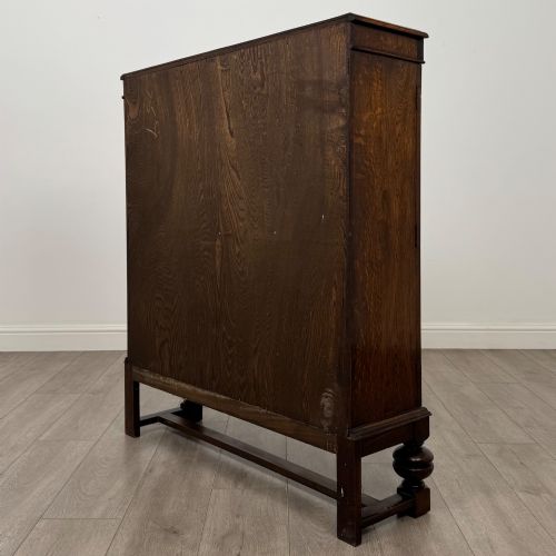 fig 6. An Edwardian dark oak display cabinet with double diamond-leaded glass doors, a carved scroll frieze along the top, interior wooden shelves, and bulbous turned legs connected by a stretcher.