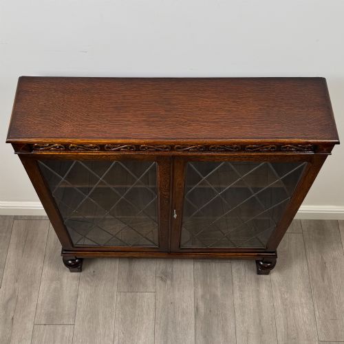 fig 9. An Edwardian dark oak display cabinet with double diamond-leaded glass doors, a carved scroll frieze along the top, interior wooden shelves, and bulbous turned legs connected by a stretcher.