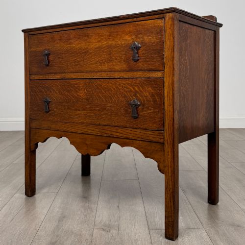 fig 1. An antique golden oak chest of drawers featuring two wide drawers with stylised dark metal handles and a scalloped lower apron.