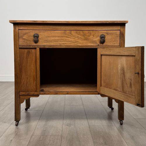 fig 1. An early 20th-century golden oak side cabinet featuring a top drawer with ornate metal drop handles above a central panelled cupboard door on square legs with castors.
