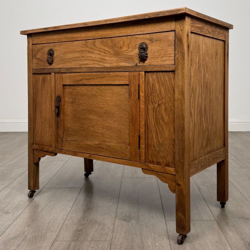 fig 2. An early 20th-century golden oak side cabinet featuring a top drawer with ornate metal drop handles above a central panelled cupboard door on square legs with castors.