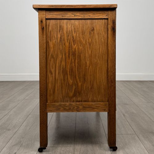 fig 3. An early 20th-century golden oak side cabinet featuring a top drawer with ornate metal drop handles above a central panelled cupboard door on square legs with castors.