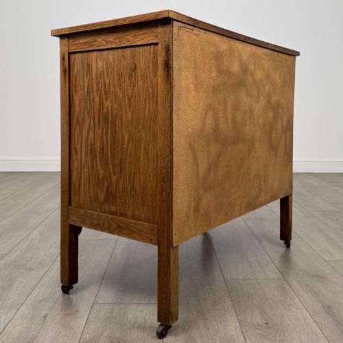 fig 4. An early 20th-century golden oak side cabinet featuring a top drawer with ornate metal drop handles above a central panelled cupboard door on square legs with castors.