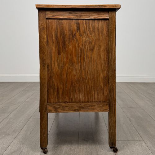 fig 7. An early 20th-century golden oak side cabinet featuring a top drawer with ornate metal drop handles above a central panelled cupboard door on square legs with castors.