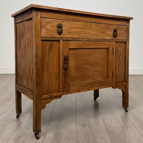 fig 8. An early 20th-century golden oak side cabinet featuring a top drawer with ornate metal drop handles above a central panelled cupboard door on square legs with castors.