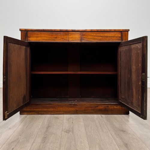 fig 1. A 19th-century Regency mahogany cupboard featuring two frieze drawers, full-length turned flanking columns, and two lower doors set with pleated gold-tone fabric panels on a solid plinth base.