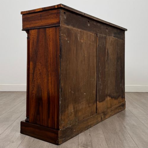 fig 4. A 19th-century Regency mahogany cupboard featuring two frieze drawers, full-length turned flanking columns, and two lower doors set with pleated gold-tone fabric panels on a solid plinth base.