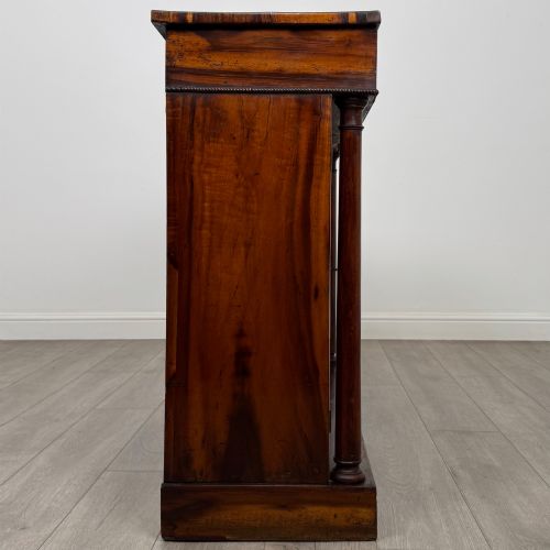 fig 7. A 19th-century Regency mahogany cupboard featuring two frieze drawers, full-length turned flanking columns, and two lower doors set with pleated gold-tone fabric panels on a solid plinth base.