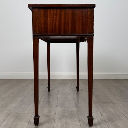 fig 6. A dark mahogany 20th-century Adam style side table featuring a central carved urn motif, fluted frieze drawers, brass ring handles, and square tapered legs ending in spade feet.