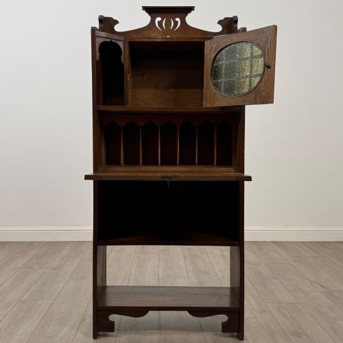 fig 1. An Arts and Crafts dark oak writing bureau featuring a top crest with a pierced heart motif, a small upper cupboard with a leaded oval window, a fall-front writing section, and two open lower shelves.