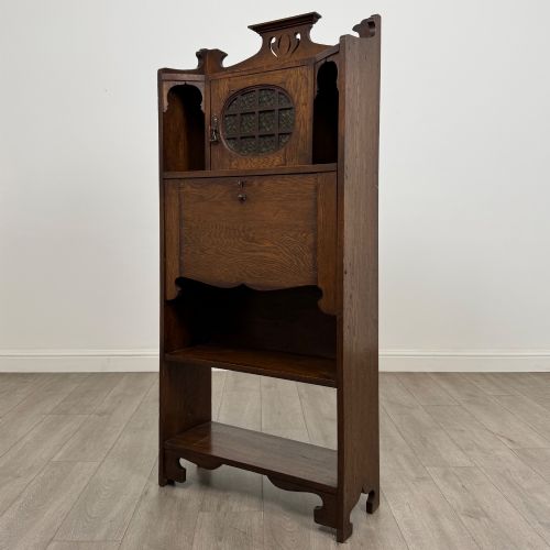 fig 2. An Arts and Crafts dark oak writing bureau featuring a top crest with a pierced heart motif, a small upper cupboard with a leaded oval window, a fall-front writing section, and two open lower shelves.