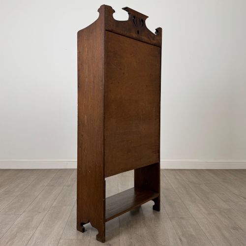 fig 4. An Arts and Crafts dark oak writing bureau featuring a top crest with a pierced heart motif, a small upper cupboard with a leaded oval window, a fall-front writing section, and two open lower shelves.