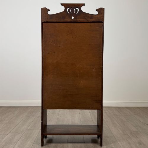 fig 5. An Arts and Crafts dark oak writing bureau featuring a top crest with a pierced heart motif, a small upper cupboard with a leaded oval window, a fall-front writing section, and two open lower shelves.