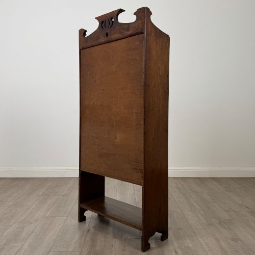 fig 6. An Arts and Crafts dark oak writing bureau featuring a top crest with a pierced heart motif, a small upper cupboard with a leaded oval window, a fall-front writing section, and two open lower shelves.
