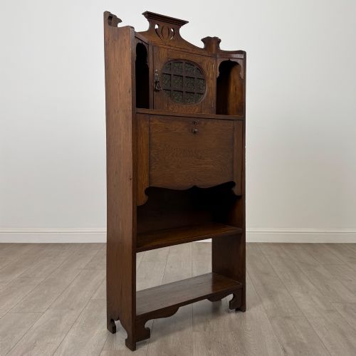 fig 8. An Arts and Crafts dark oak writing bureau featuring a top crest with a pierced heart motif, a small upper cupboard with a leaded oval window, a fall-front writing section, and two open lower shelves.