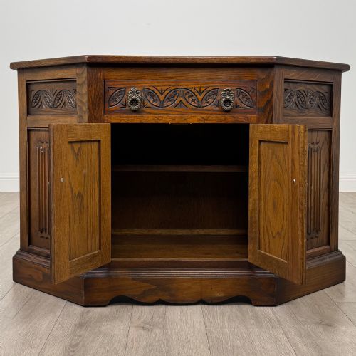 fig 1. A dark oak low cupboard featuring canted corners, carved linenfold door panels, an upper drawer with carved foliage, and brass hinges.