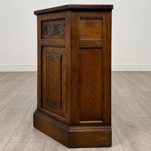 fig 3. A dark oak low cupboard featuring canted corners, carved linenfold door panels, an upper drawer with carved foliage, and brass hinges.