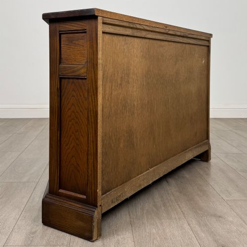fig 4. A dark oak low cupboard featuring canted corners, carved linenfold door panels, an upper drawer with carved foliage, and brass hinges.