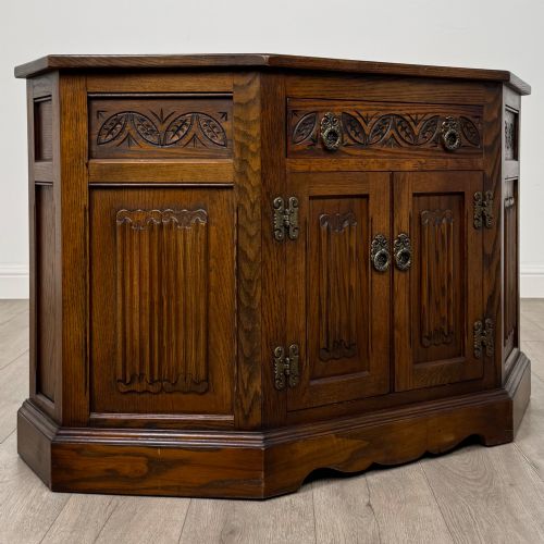 fig 8. A dark oak low cupboard featuring canted corners, carved linenfold door panels, an upper drawer with carved foliage, and brass hinges.