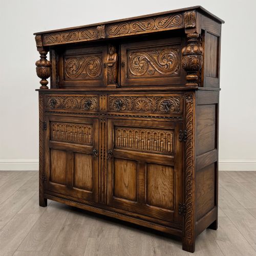fig 2. A dark carved oak court cupboard featuring two top doors with relief carving, bulbous turned supports, two drawers, and two panelled cupboard doors below.