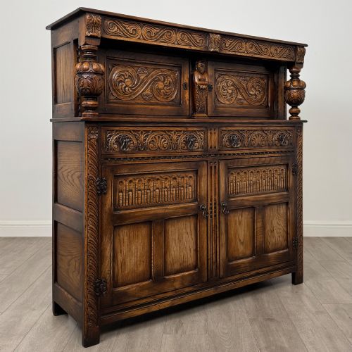 fig 8. A dark carved oak court cupboard featuring two top doors with relief carving, bulbous turned supports, two drawers, and two panelled cupboard doors below.