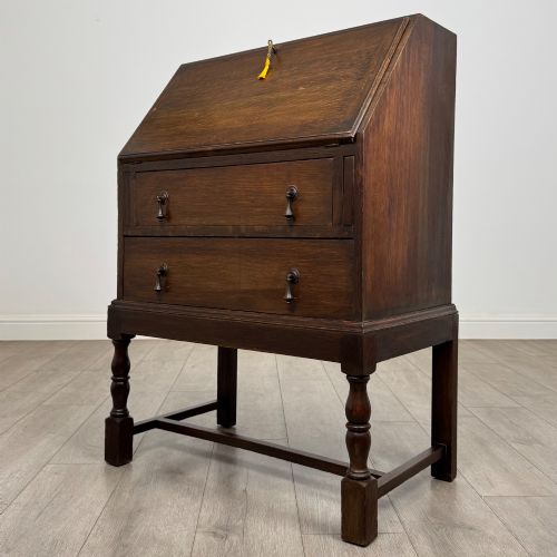 fig 2. An Edwardian dark oak writing bureau featuring a sloped drop-down lid, two lower drawers with teardrop handles, and turned legs connected by stretchers.