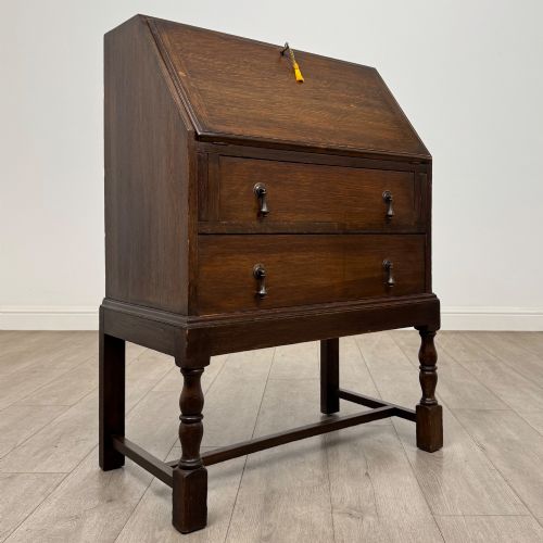 fig 8. An Edwardian dark oak writing bureau featuring a sloped drop-down lid, two lower drawers with teardrop handles, and turned legs connected by stretchers.