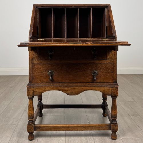 fig 1. An Edwardian golden oak writing bureau featuring a slant-front writing flap above two drawers with brass teardrop handles, supported on turned legs with a perimeter stretcher.