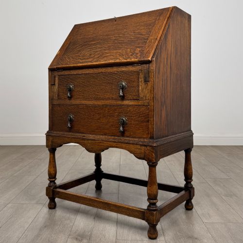 fig 2. An Edwardian golden oak writing bureau featuring a slant-front writing flap above two drawers with brass teardrop handles, supported on turned legs with a perimeter stretcher.