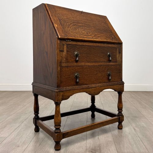 fig 8. An Edwardian golden oak writing bureau featuring a slant-front writing flap above two drawers with brass teardrop handles, supported on turned legs with a perimeter stretcher.