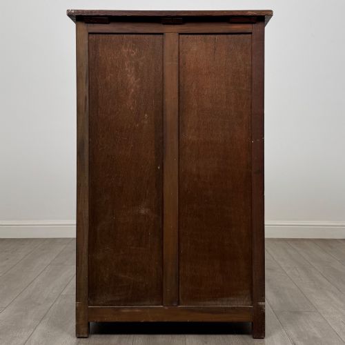 fig 5. A dark oak cabinet featuring carved top frieze detailing, two arched fielded panel doors with exposed iron hinges, and two lower drawers with turned wooden knobs.