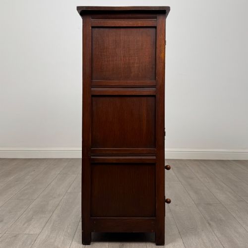 fig 7. A dark oak cabinet featuring carved top frieze detailing, two arched fielded panel doors with exposed iron hinges, and two lower drawers with turned wooden knobs.