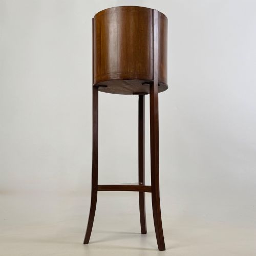 fig 1. A tall Edwardian mahogany plant stand featuring a cylindrical top bucket with subtle line inlay, supported by three elongated curved legs and a triangular lower shelf.