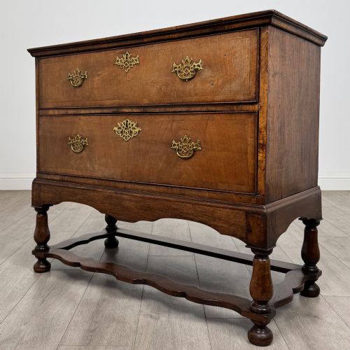 fig 1. An antique 19th-century William and Mary style walnut chest of drawers featuring two deep drawers with ornate brass drop handles, raised on turned legs connected by a wavy stretcher base.