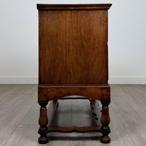 fig 6. An antique 19th-century William and Mary style walnut chest of drawers featuring two deep drawers with ornate brass drop handles, raised on turned legs connected by a wavy stretcher base.